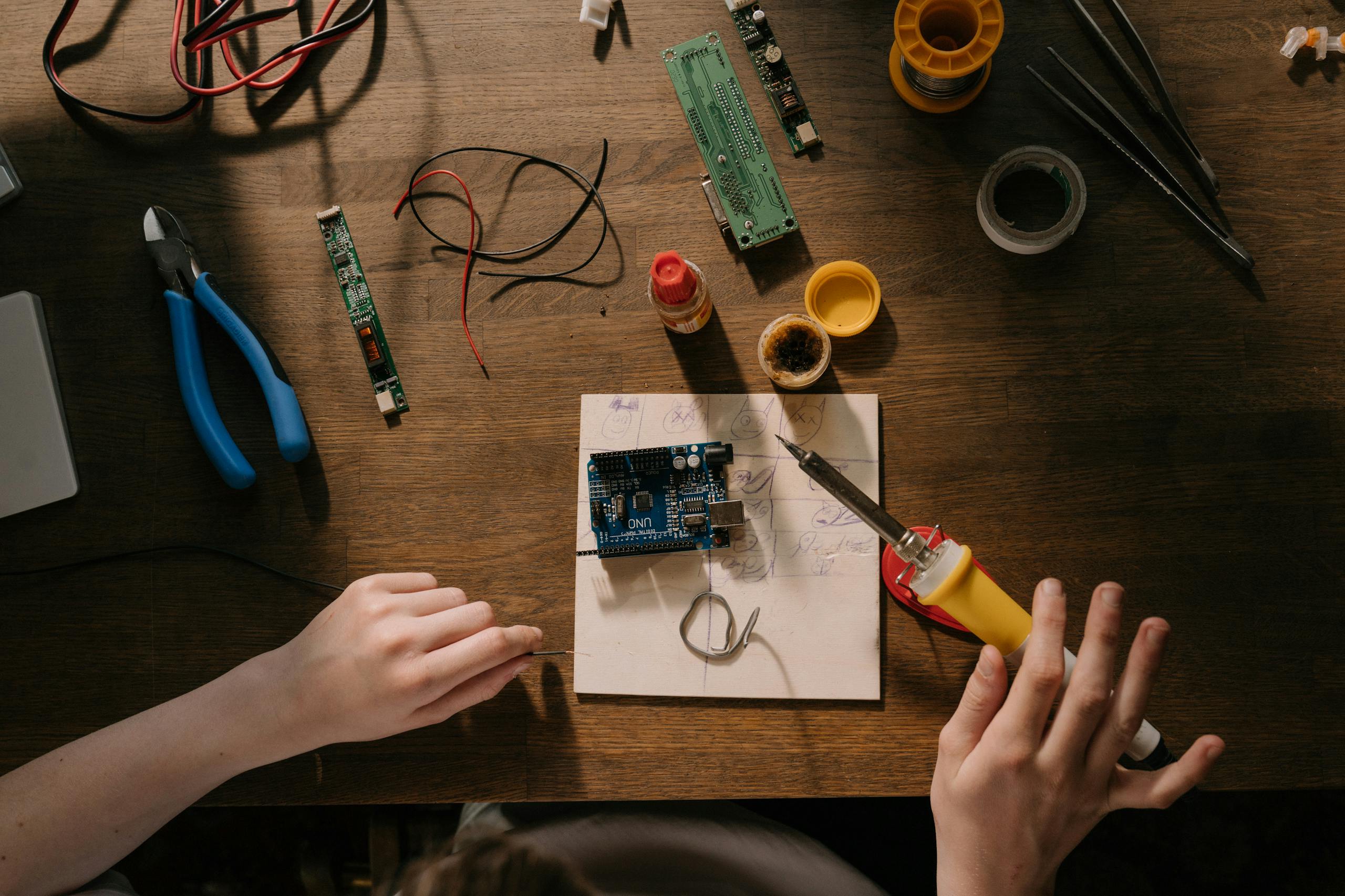 Top view of a soldering setup with circuit board, tools, and components on a wooden desk.