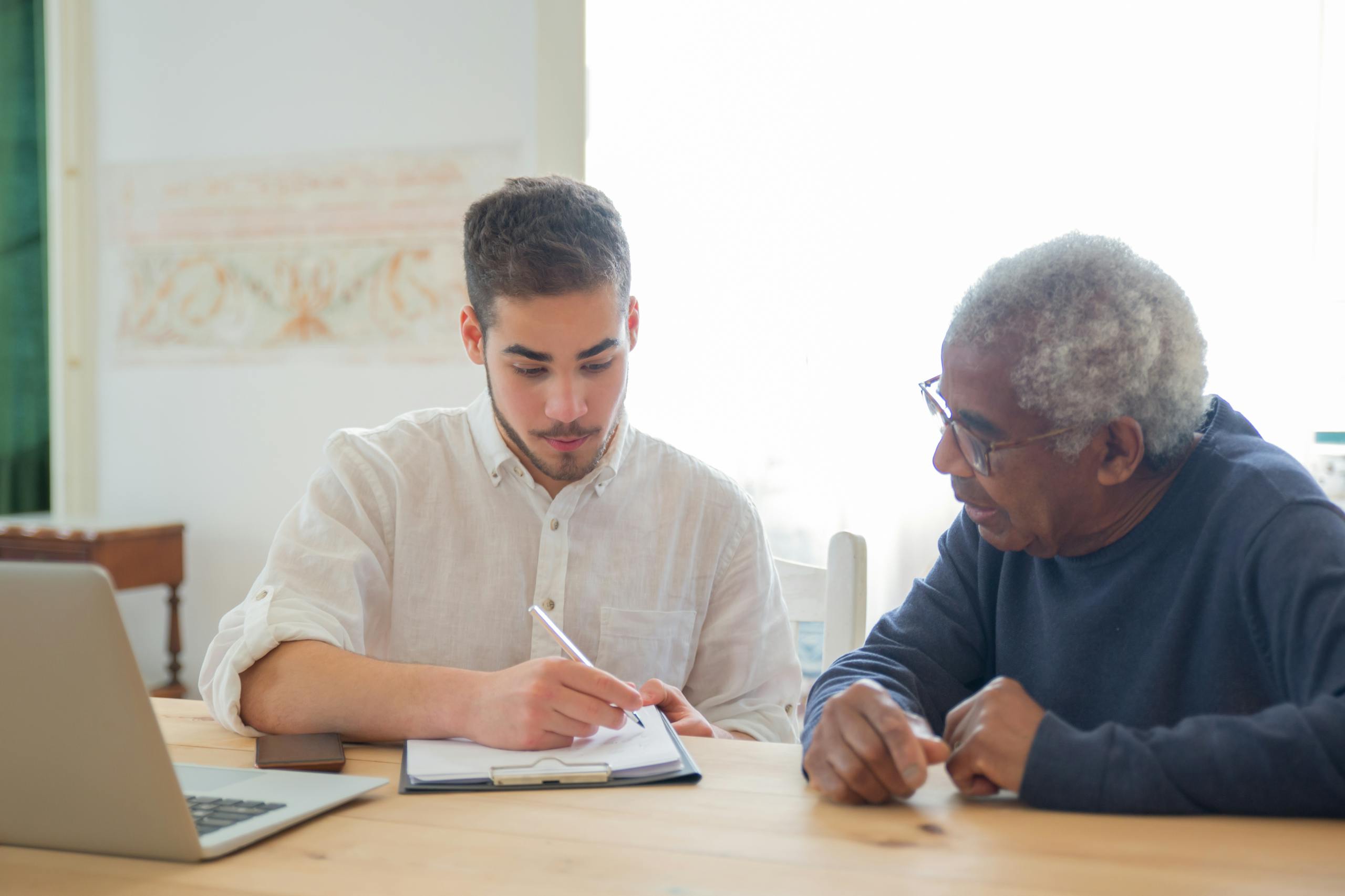 Young man helps elderly man with documents at a desk indoors, promoting collaboration.